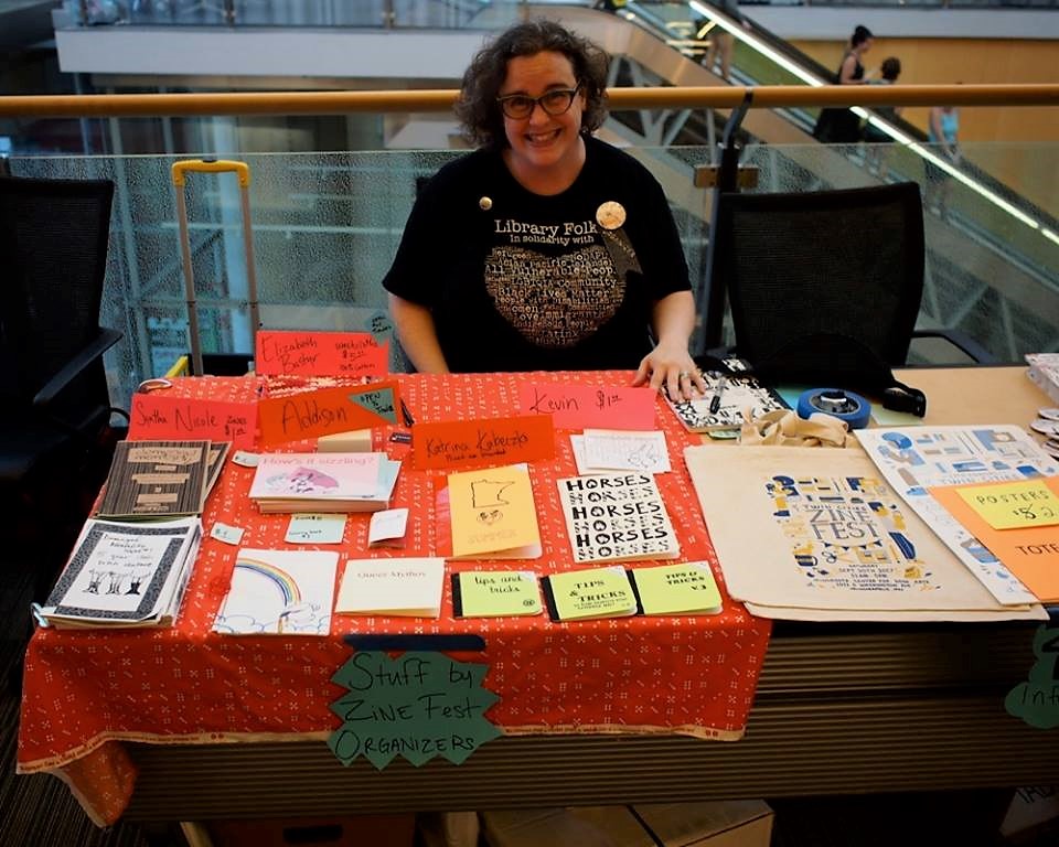 Violet sitting at a table full of zines at the 2018 Twin Cities Zine Fest with a big grin.