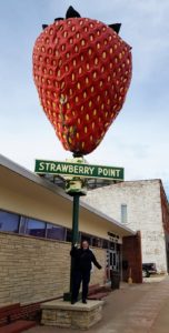 Violet standing underneath a ten foot strawberry statue on top of a twelve foot pole. Underneath the strawberry is a sign reading Strawberry Point, Iowa